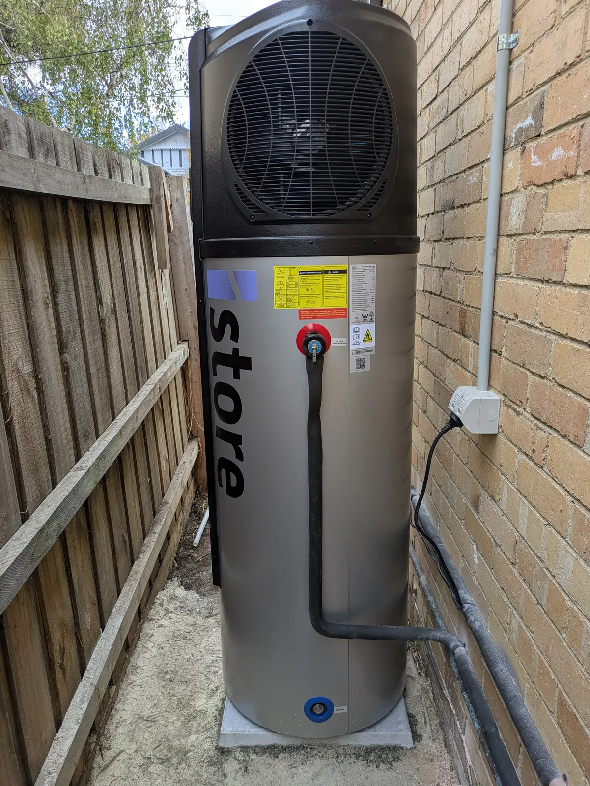 An iStore heat pump installed in a narrow side passage between a brick wall and a paling fence