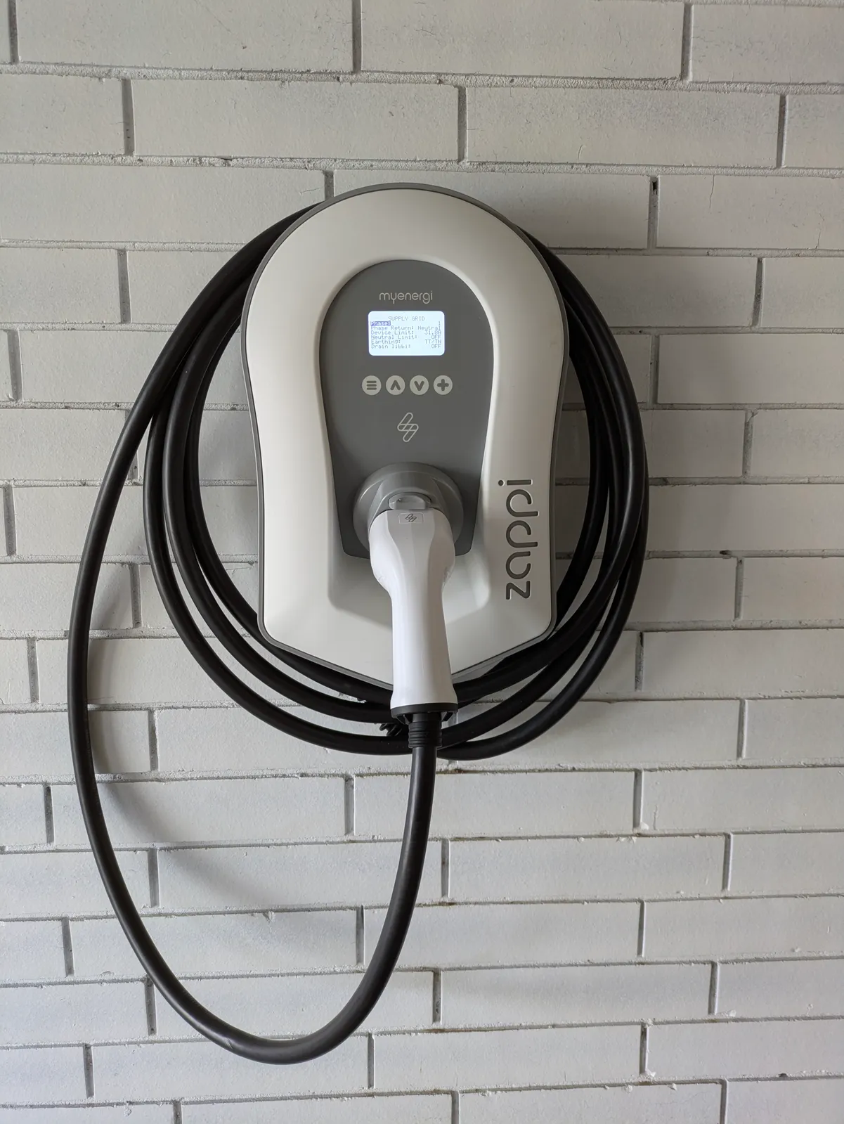 A myenergi Zappi on a white brick wall, display lit and the tethered cable coiled